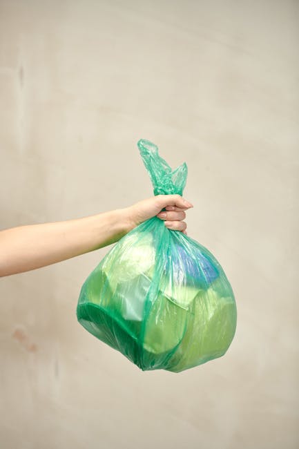 A person’s arm is extended across the frame, holding a green and blue plastic rubbish bag by its twisted, knotted top against a neutral beige background. The bag appears to be medium-sized, semi-tra