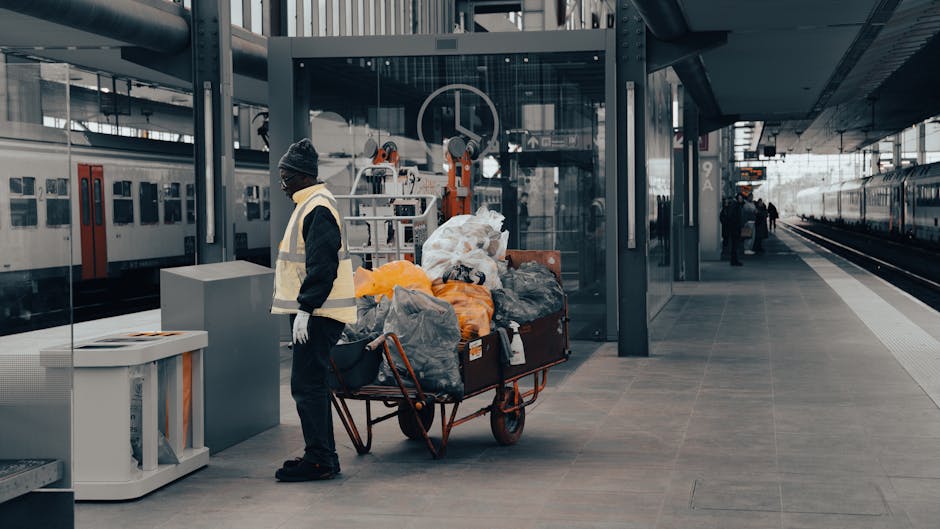 An outdoor scene in an urban area showing a large pile of mixed domestic waste and rubbish overflowing from a metal recycling bin with a blue lid, situated on a paved sidewalk. The waste includes crum